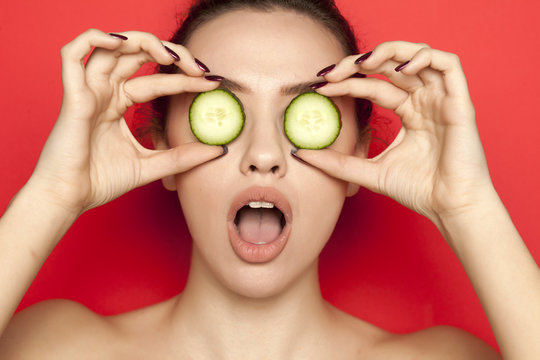 Sexy young woman posing with slices of cucumber on her face on red background