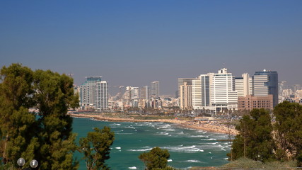 Widok na zatokę Morza śródziemnego w Tel Awiwie, Izrael, z piaszczystą plażą i nowoczesnymi wysokimi budynkami na nabrzeżu, na pierwszym planie drzewa  w parku w Hajfie, fale na szmaragdowej wodzie