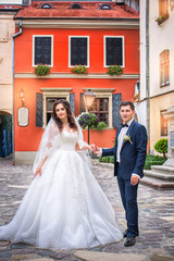 Fototapeta premium Beautiful bride and stylish bridegroom holding hands on the background of an orange building