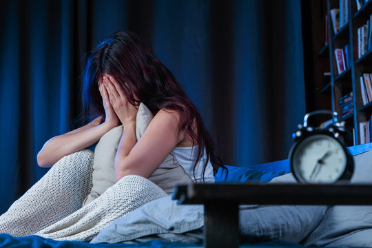 Image of unhappy woman with insomnia sitting on bed next to alarm clock
