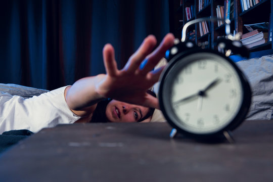 Image of dissatisfied woman with insomnia stretching arm to alarm clock at night