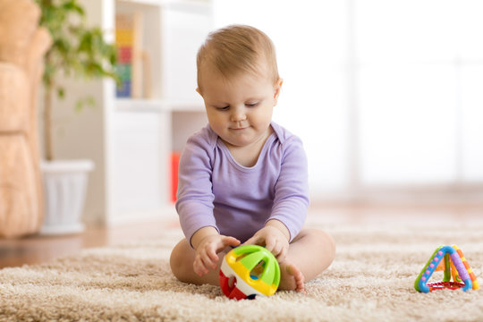 Cute baby girl playing with colorful toys sitting on carpet in white sunny bedroom. Child with educational toy. Early development.