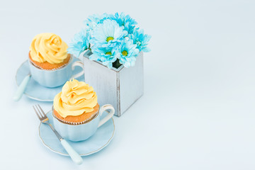 Cupcake with tender yellow cream decoration and bouquet of blue chrysanthemum in retro shabby chic vase on blue pastel background.