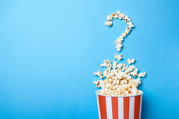 Concept of which film to choose. Spilled popcorn in the form of question mark and a paper bucket in red strip on blue background. Copy space for text - obrazy, fototapety, plakaty