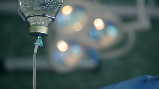 A soft focus macro of a transparent drop counter with plastic tubes and special liquid. Several lamps are seen in the surgery background.