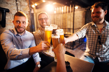 A group of young cheerful men clinking glasses with a beer in the sunny pub after work.