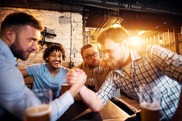 Group of joyful happy men having fun in the pub. Two friends arm wrestling while other supporting.