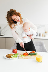 Portrait of young mother standing and holding her little baby while talking on her cellphone and cooking on kitchen isolated