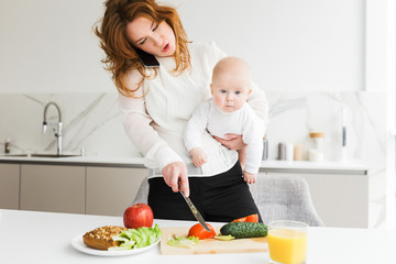 Close up photo of young mother standing and holding her little baby while talking on her cellphone and cooking on kitchen isolated