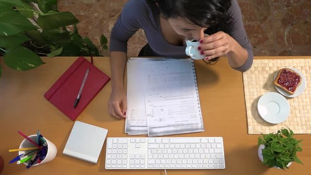 Caucasian woman on desk writing on computer and having breakfast at home. Active businesswoman working at office proofreading draft text, drinking coffee and eating bread and jam. Busy lifestyle