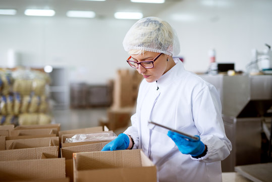 Young gorgeous concerned female worker in sterile cloths using a tablet to check correction of inventory within boxes in factory storage room.