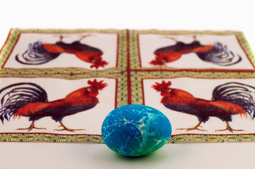 A painted Easter egg with an abstract pattern in blue, green tones with a divorce on the background of a napkin with a picture of a cock.
