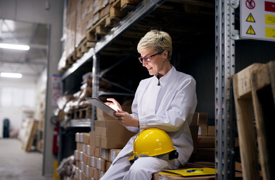 Young Female Dedicated Engineer Is Sitting On Stacks Of Boxes And Using Her Tablet While Having A Helmet On Her Knee In Storage Cargo Area.