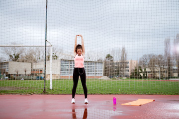 Obraz premium Motivated attractive young fitness girl stretching while holding hands above head outside near the soccer field.