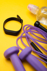 Sports accessories - jump rope, dumbbells, fitness bracelet and water. Yellow background.