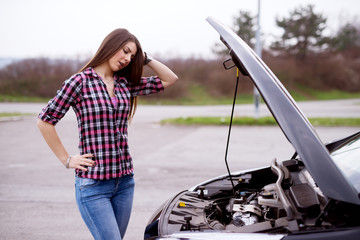 Side view of a young worried beautiful girl is looking under the hood of her car and holding her head in despair.
