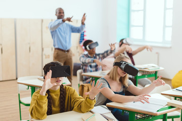 High school teenage students using virtual reality headsets and gesturing teacher standing behind in classroom - Powered by Adobe