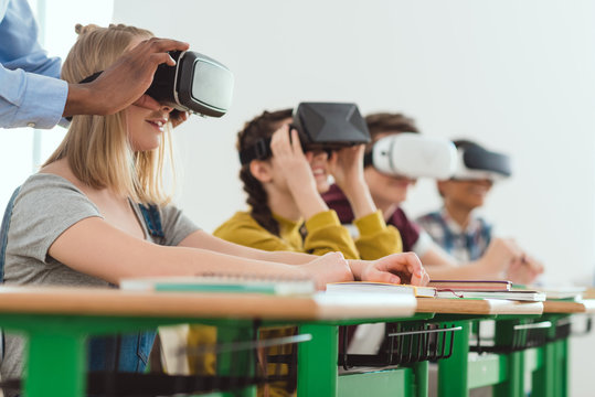 Cropped image of african american teacher adjusting virtual reality headset to teenage schoolgirl with classmates sitting behind