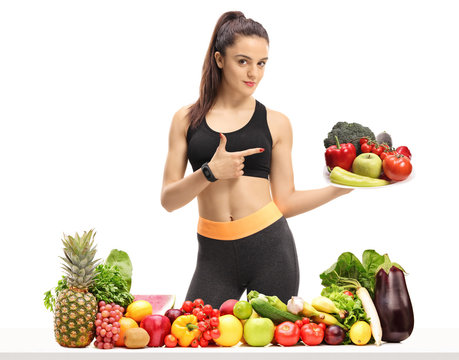 Fitness girl behind a table with fruit and vegetables holding a plate and pointing