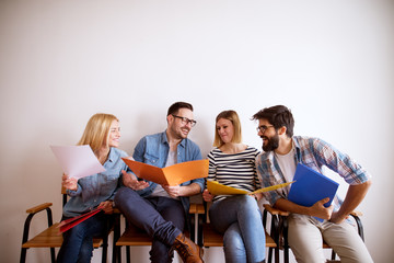 Group of young joyful satisfied friends holding colourful folder while sitting in a chair sharing notes and laughing in the waiting room for an exam.