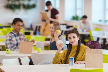 smiling teen schoolgirl sitting at school cafeteria with classmates and looking at camera
