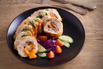 Chicken wrapped with ham and cheddar cheese; vegetables in black plate on wooden table. horizontal