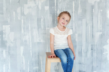 Sweet happy little girl in blank white t-shirt sitting on chair against grey textured wall background