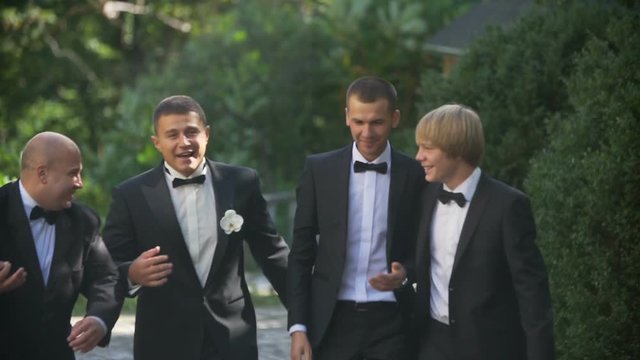 Wedding concept. The young handsome groom is happily talking with his three best man while walking along the garden.