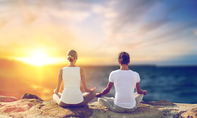Fototapeta premium yoga , mindfulness, harmony and people concept - happy couple meditating in lotus pose outdoors over sea background