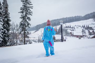 Girl in blue, pink unicorn pijama kigurumi outdoor in front of the wood houses on the ski report in snow mountains.