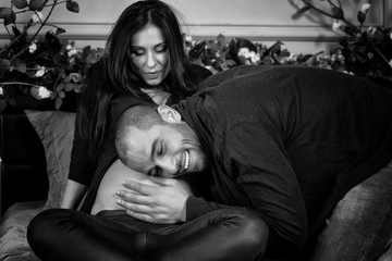 Black and white photo of enamored international couple, man listening his pregnant wife's belly while sitting on the gray cozy bed