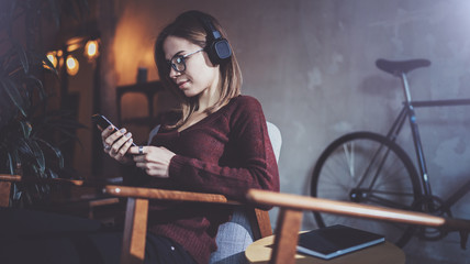 Fototapeta na wymiar Young attractive hipster girl wearing eye glasses and casual clothes listening music in headphones on smartphone at modern place.