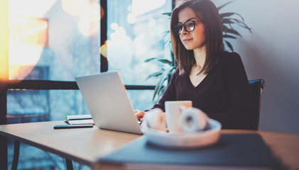 Young smiling girl wearing eye glasses working on laptop at her workplace at night office.Horizontal.Blurred background.Flares.
