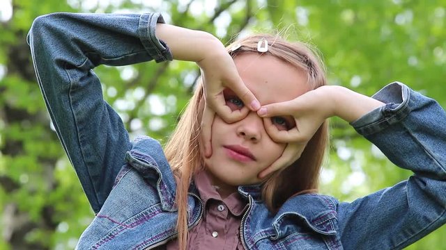 Cute caucasian teenager girl making silly faces and laughing. Closeup portrait of funny pretty teenage kid forming glasses for eyes with her fingers outdoors.