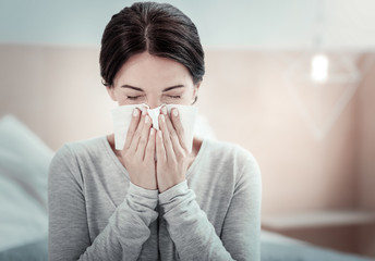 Home treatment. Ill exhausted unhappy woman sitting in the bright room holding a napkin and...