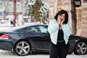 Fototapeta na wymiar Stylish african american plus size model with mobile phone at hand at winter snowy day against black car.