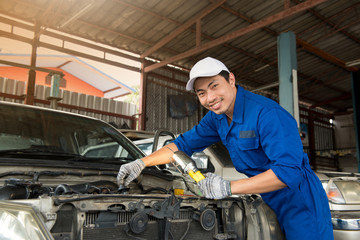 Smiling asian mechanic in blue uniform and white hat at the repair garage, holding lamp look at the camera