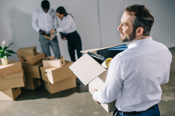 back view of businessman holding cardboard box with office supplies while colleagues unpacking boxes in new office