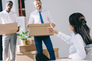 Fototapeta premium businesswoman showing thumb up to male colleagues holding cardboard boxes in new office