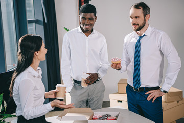 smiling multiethnic coworkers drinking coffee to go and eating donuts in new office