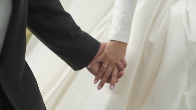 Close-up shot of the newlywed cou ple is softly holdign hands while walking. No face.