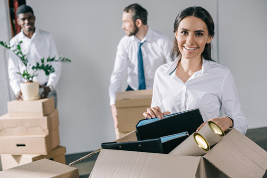 happy young businesswoman unpacking box with office supplies and smiling at camera while male colleagues standing behind in new office - Powered by Adobe
