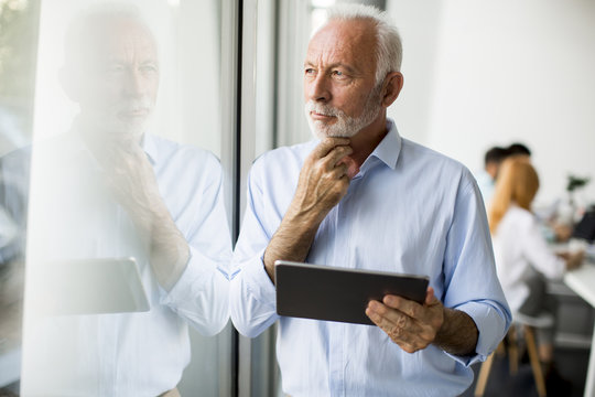 Senior Businessman Standing By Window With Digital Tablet In His Hand