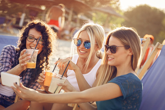 Group of young casual female friends sitting on beach on sunbeds,hangout and toasting with drink.