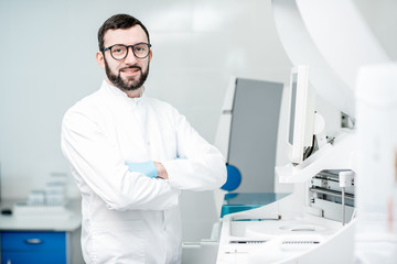 Portrait of a male laboratory assistant in uniform standing near the medical analizer machine indoors