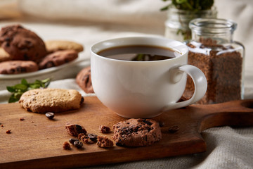 Morning coffee in white cup, chocolate chips cookies on cutting board close-up, selective focus