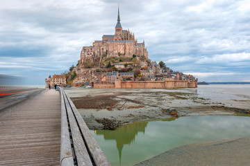 Beautiful famous Mont Saint Michel with bridge and reflection in the canal at low tide in the cloudy morning, Normandy, France