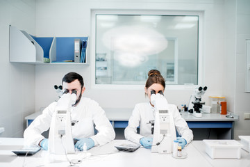 Naklejka premium Couple of medics in uniform working with microscope making analysis at the laboratory office