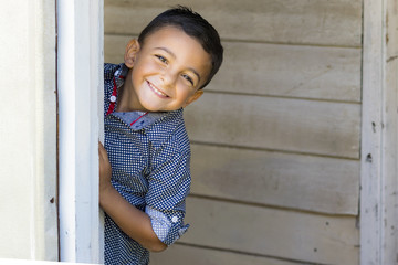 Cute little boy hiding and playing behind a door.  Outdoor portrait of a smiling little boy in blue checkered shirt.