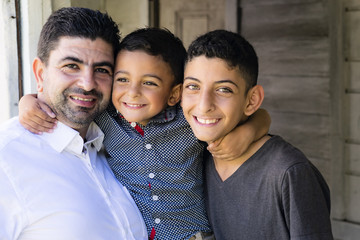 Outdoor portrait of a father and his sons. Father and older brother smiling and hugging cute little boy.